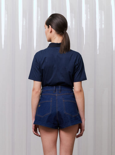 Person wearing a navy shirt and denim shorts against a white curtain background