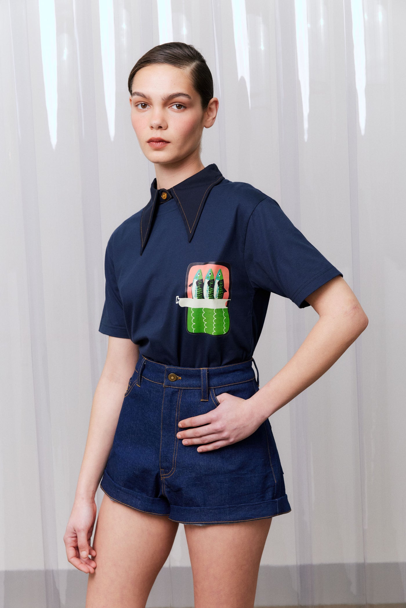Model wearing a navy blue shirt with a sardine design and denim shorts against a white curtain background