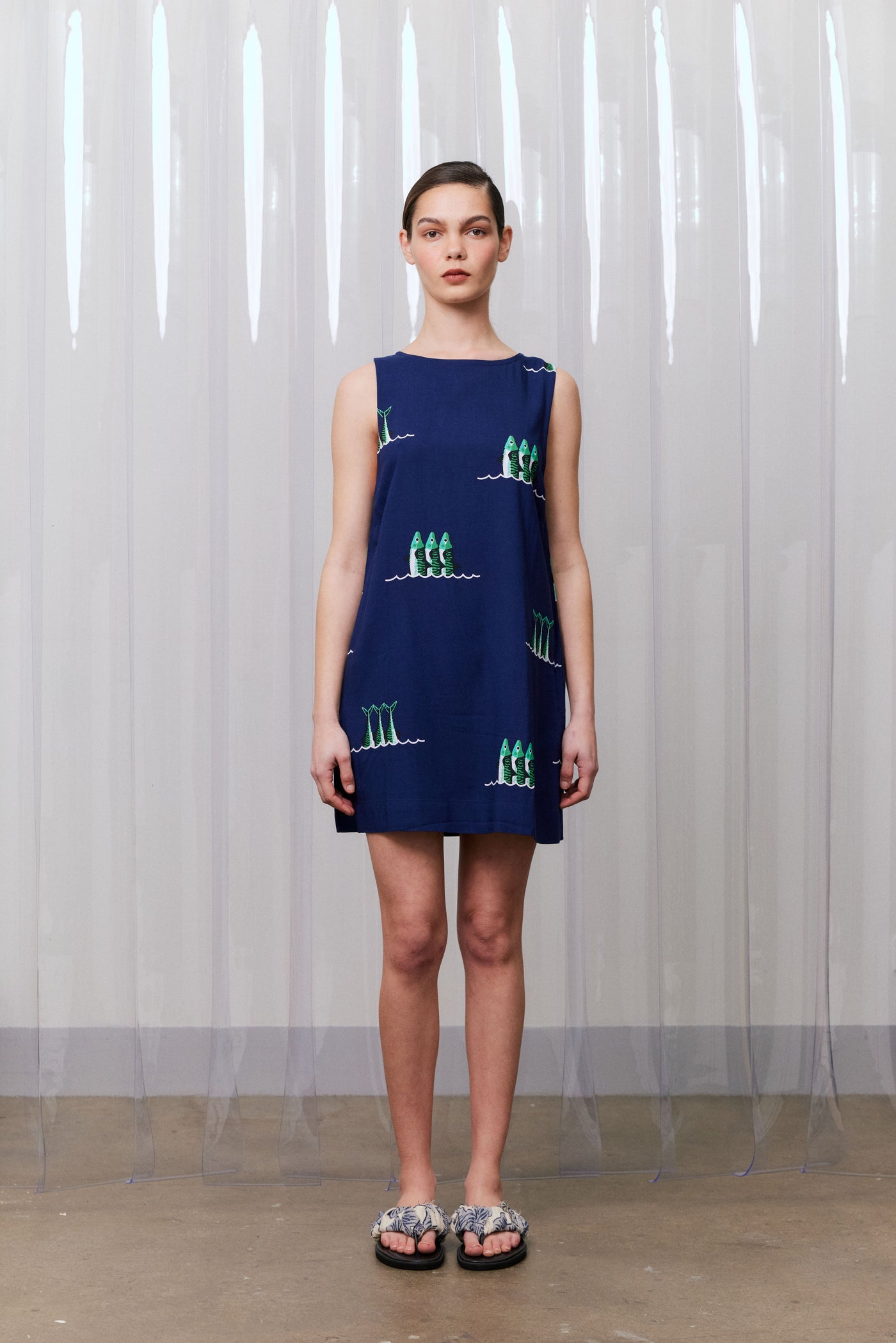 Woman wearing a blue sleeveless dress with green patterns against a white curtain background
