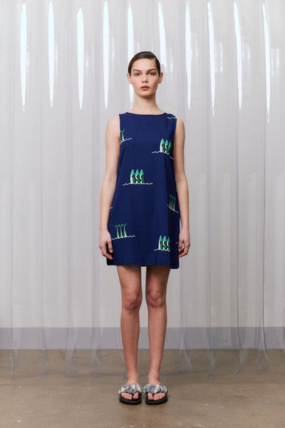 Woman wearing a blue sleeveless dress with green patterns against a white curtain background