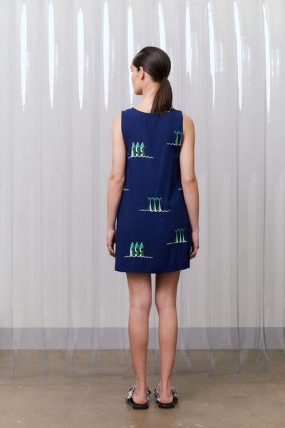 Woman wearing a navy blue dress with green patterns against a white curtain background
