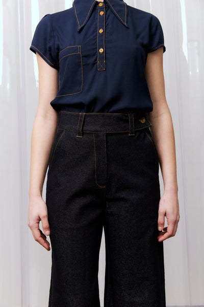 Person wearing a navy blue shirt with short sleeves and dark jeans against a white curtain background