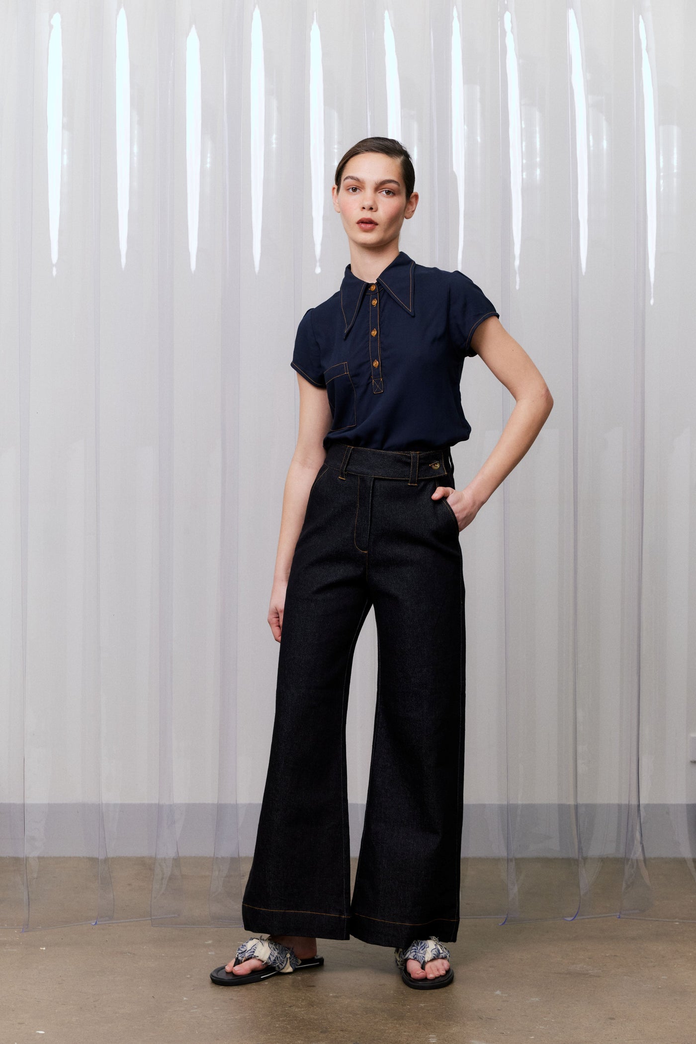 Person wearing a navy blue shirt and black jeans standing against a white curtain.
