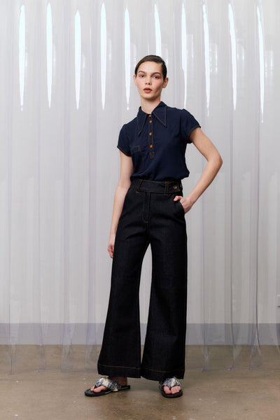Person wearing a navy blue shirt and black jeans standing against a white curtain.