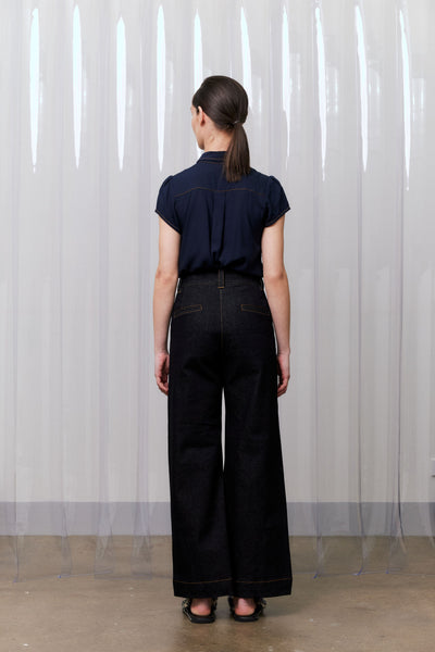 Person wearing a navy shirt and black jeans standing against a white curtain.