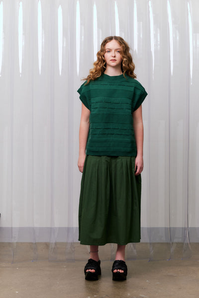 Woman wearing a green knitted vest over a khaki green dress standing against a white curtain.