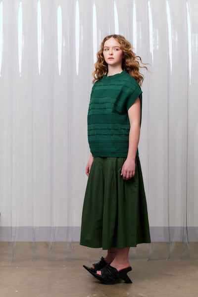 Woman wearing a green knitted vest over a khaki green dress standing against a white curtain.