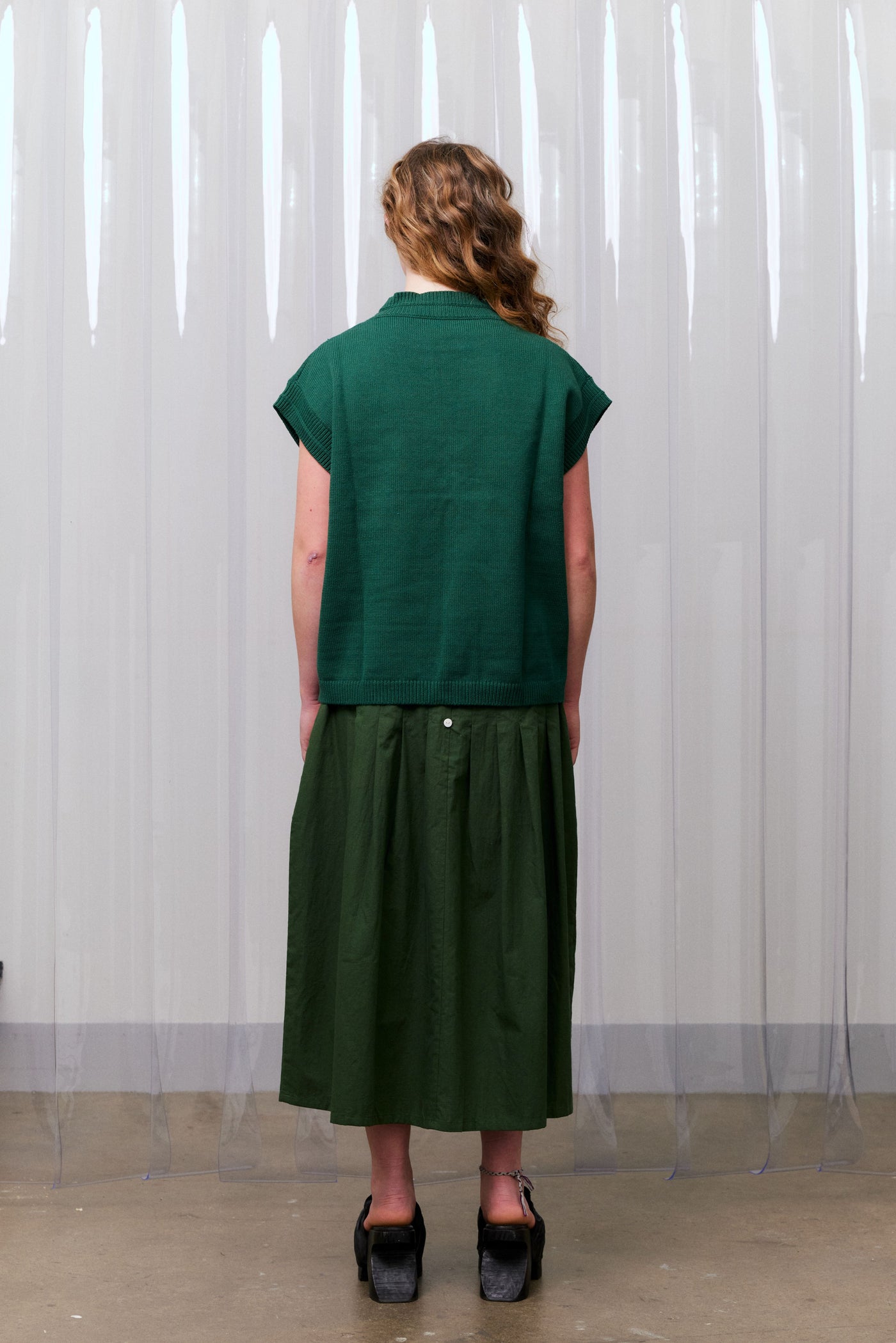 Woman wearing a green knitted vest over a khaki green dress standing against a white curtain facing away from the camera.