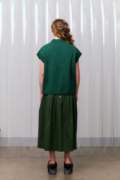 Woman wearing a green knitted vest over a khaki green dress standing against a white curtain facing away from the camera.