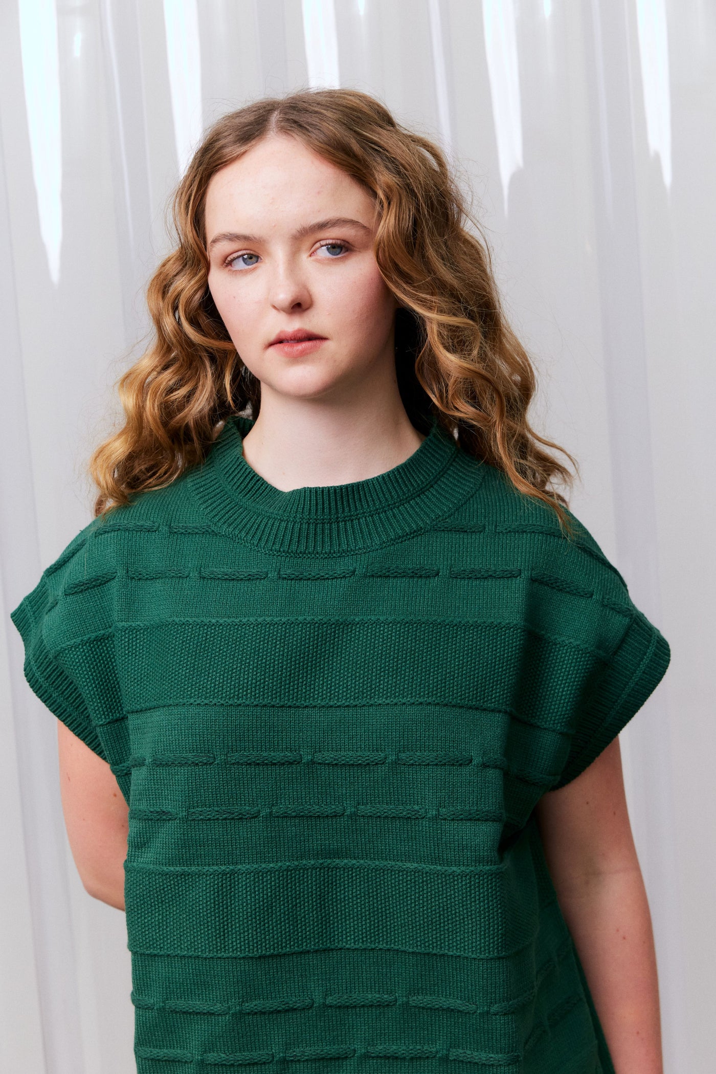 Woman wearing a green knitted vest against a white curtain background