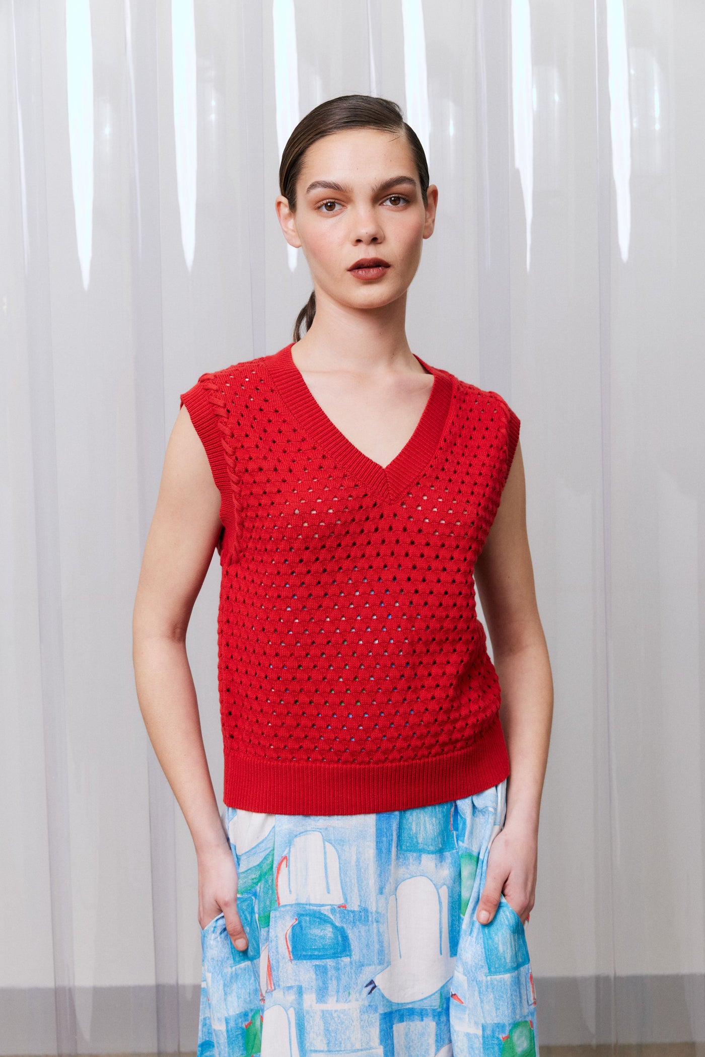 Woman wearing a red sleeveless top with a patterned skirt against a white curtain background