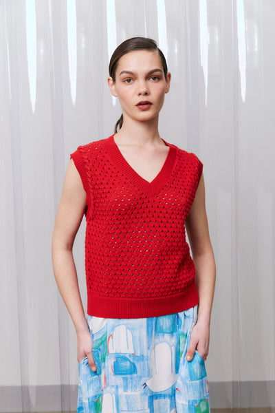 Woman wearing a red sleeveless top with a patterned skirt against a white curtain background