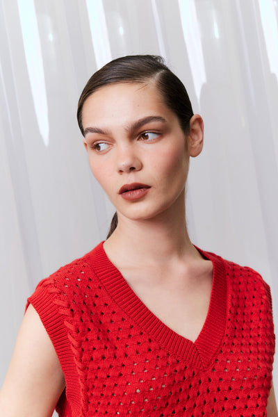 Woman wearing a red sleeveless knitted top against a white curtain background