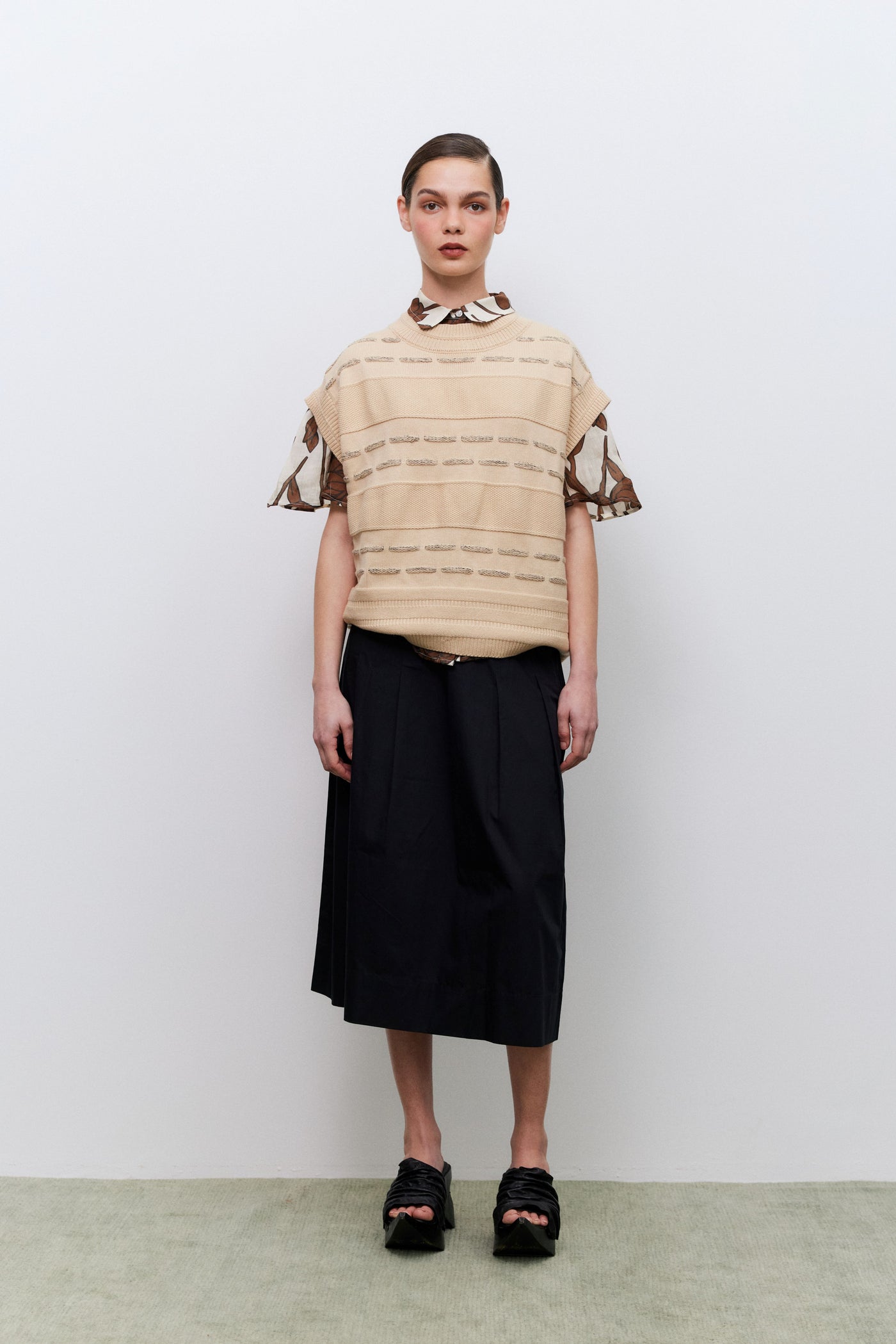 Person wearing a beige striped vest over a patterned shirt and black skirt against a white background