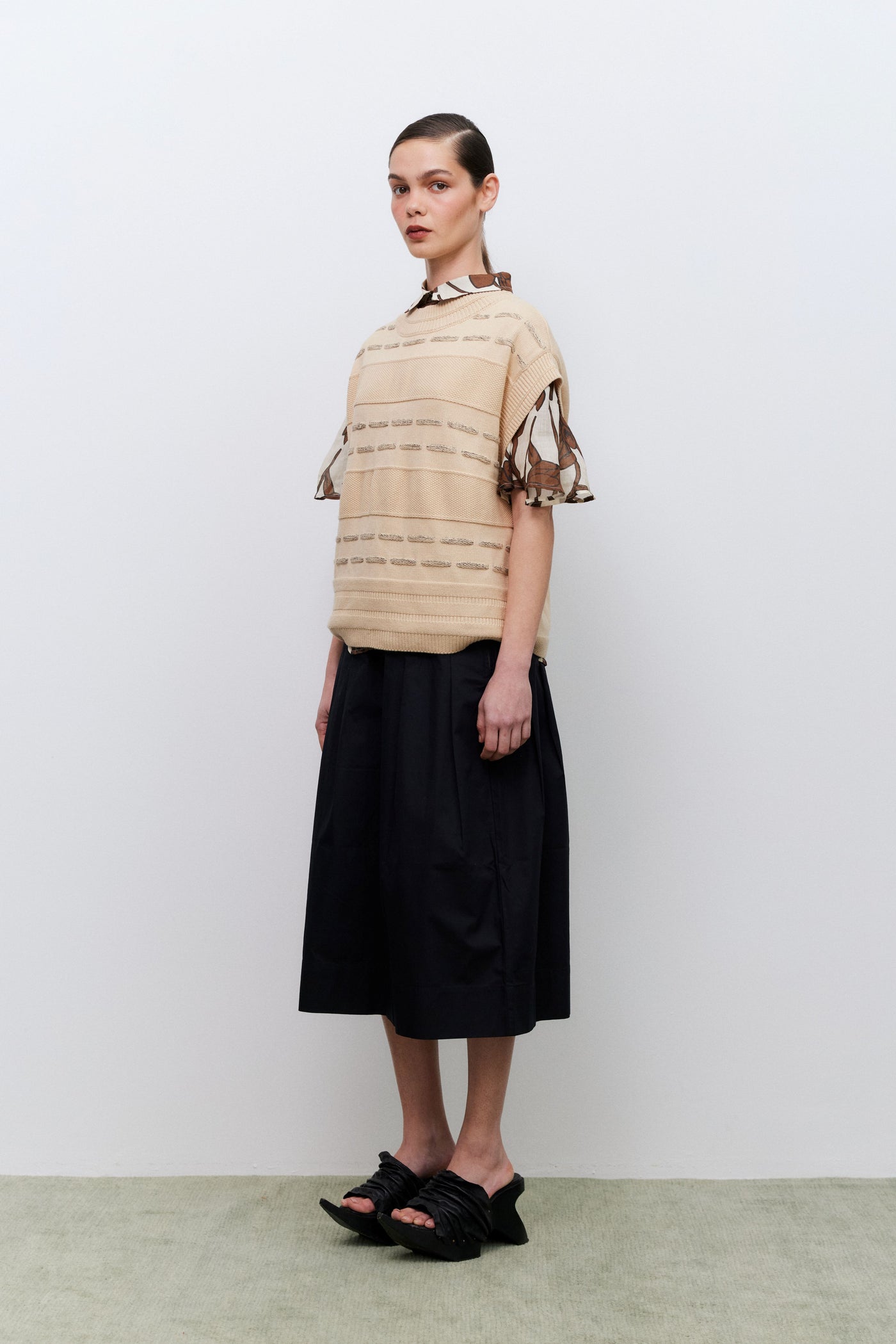 Model wearing a beige vest over a patterned top with black skirt against a white background
