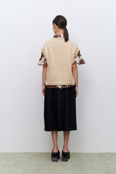 Woman wearing a beige vest over a shirt with patterned sleeves and a black skirt against a white background facing away from the camera