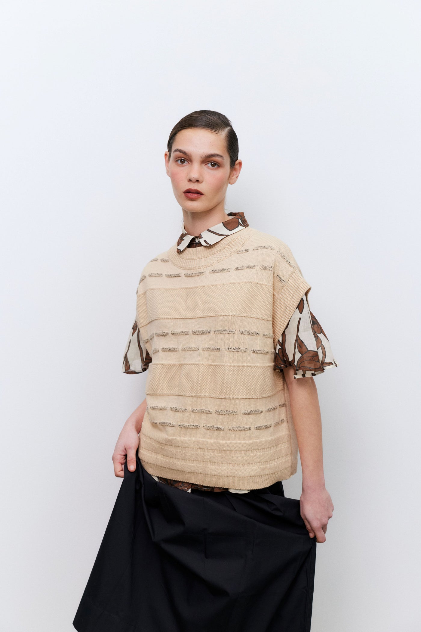 Model wearing a beige vest over a patterned top with black skirt against a white background