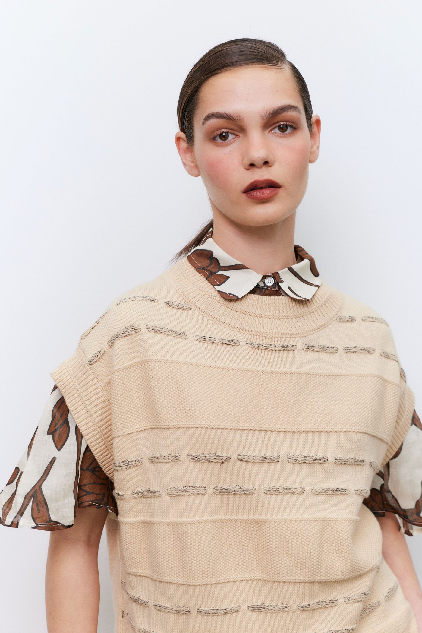 Woman wearing a beige knitted vest over a shirt with patterned sleeves and collar against a white background