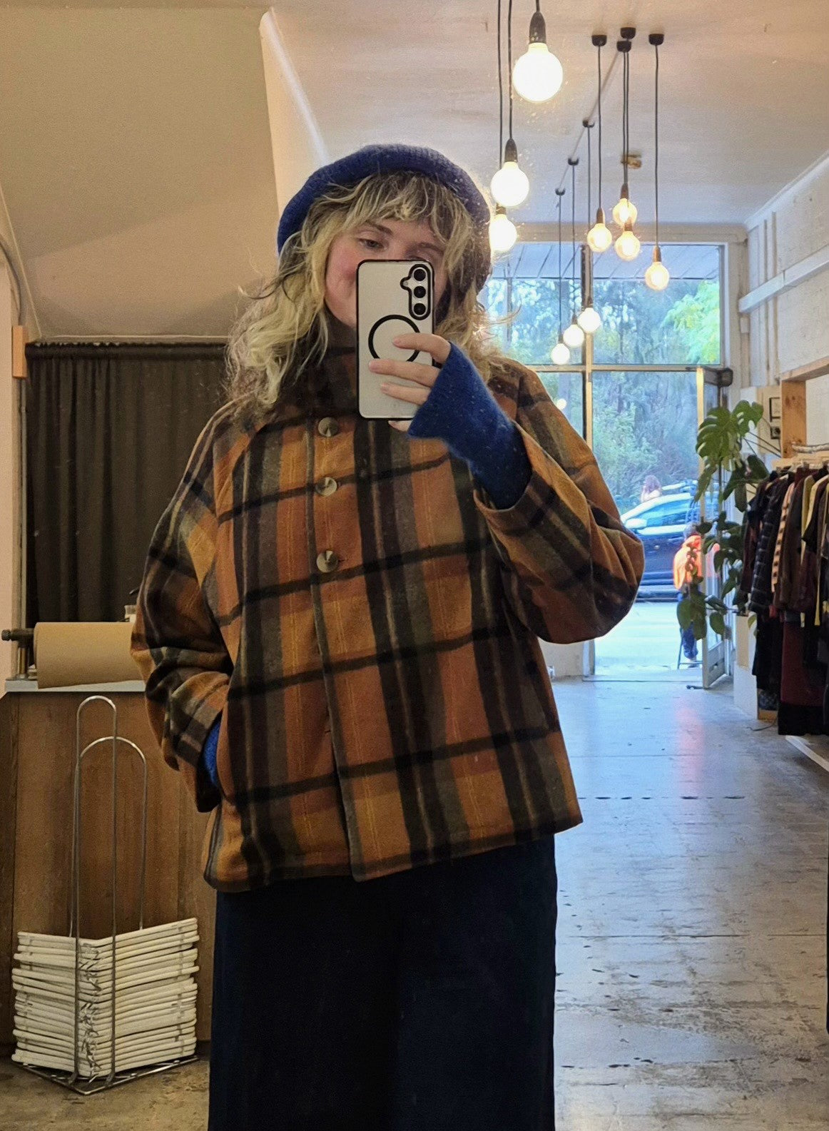 Person wearing a plaid jacket and blue beanie taking a mirror selfie in a store.