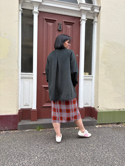 Person wearing a dark green coat and plaid skirt standing in front of a red door.