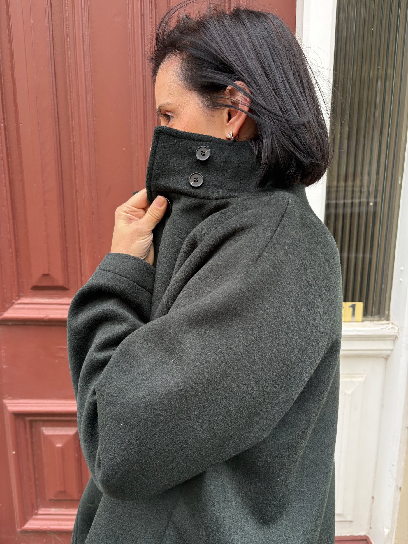 Woman wearing a dark green coat with a high collar, standing in front of a red door.