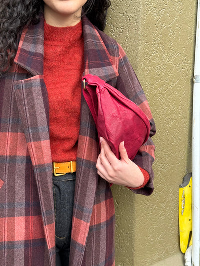 Person wearing a red sweater and plaid coat holding a red bag against a textured wall.