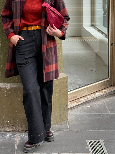 Person wearing a red sweater, plaid jacket, and dark pants standing against a wall.