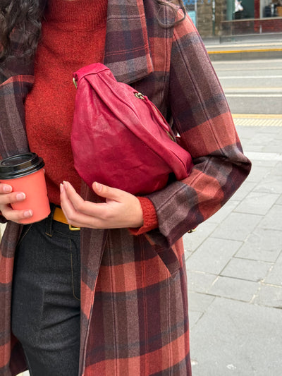 Person wearing a red sweater and plaid coat holding a red bag and a coffee cup on a street.