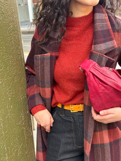 Person wearing a red sweater, plaid coat, and holding a red bag against a green wall.
