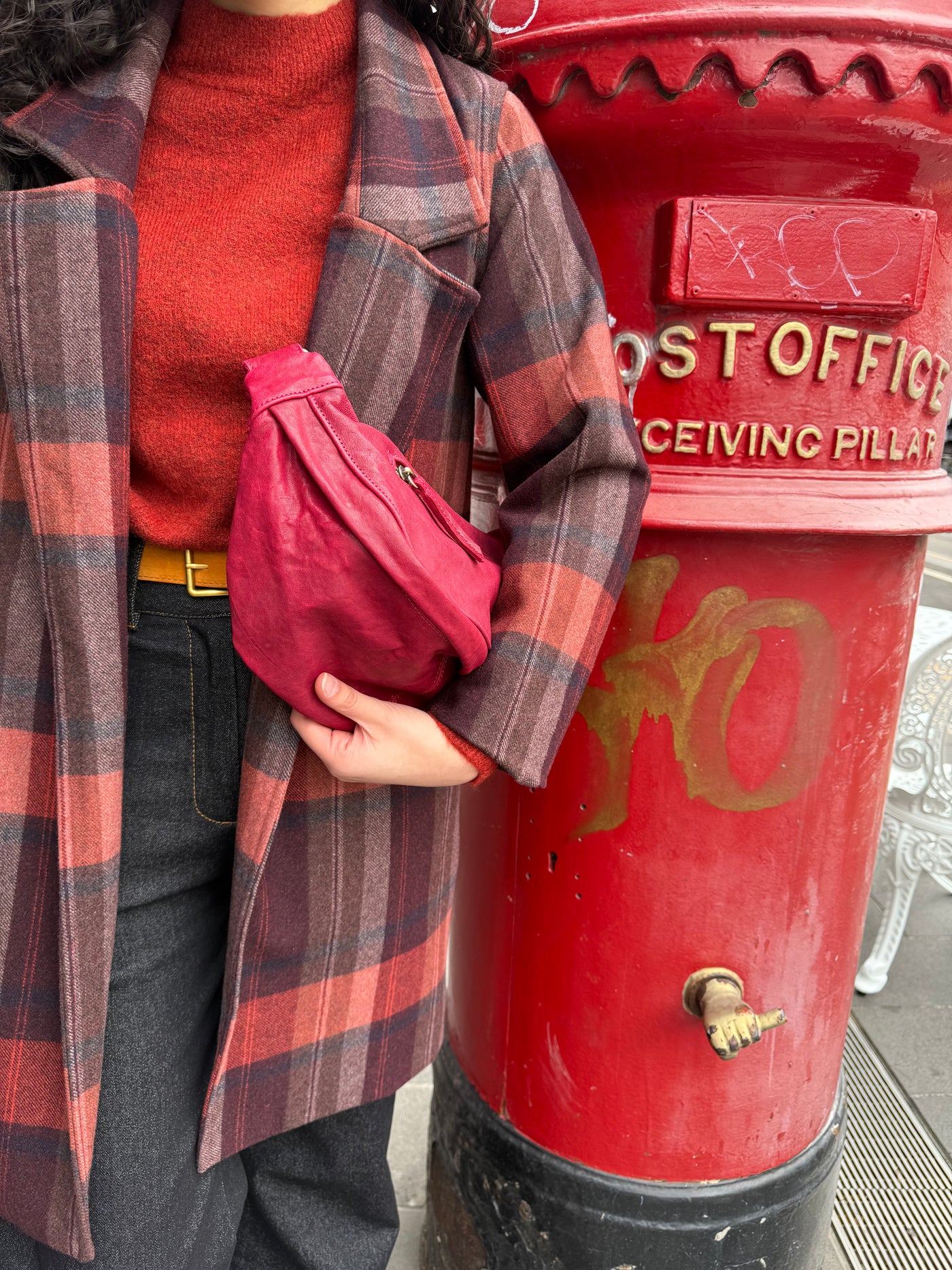 Person in red sweater and plaid coat holding a red bag next to a red post office box.