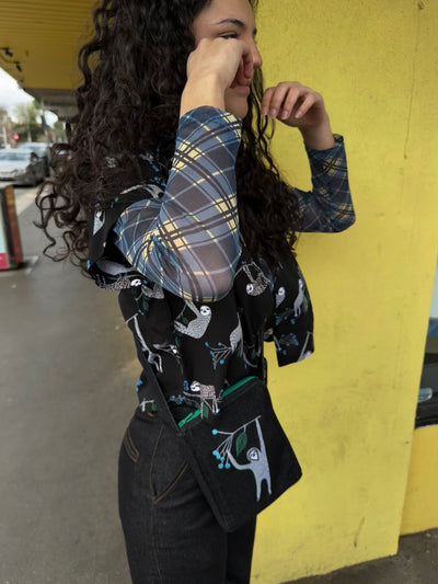 Person wearing a plaid shirt with a sloth over shirt and matching bag, standing against a yellow wall