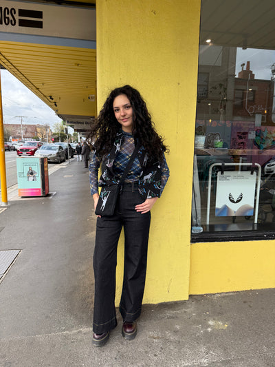 Woman standing on a sidewalk next to a yellow building wearing a mesh check top, black jeans and a satchel