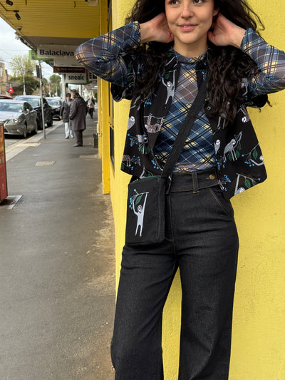 Person wearing a plaid shirt with sloth print shirt, matching bag and black pants against a yellow wall.