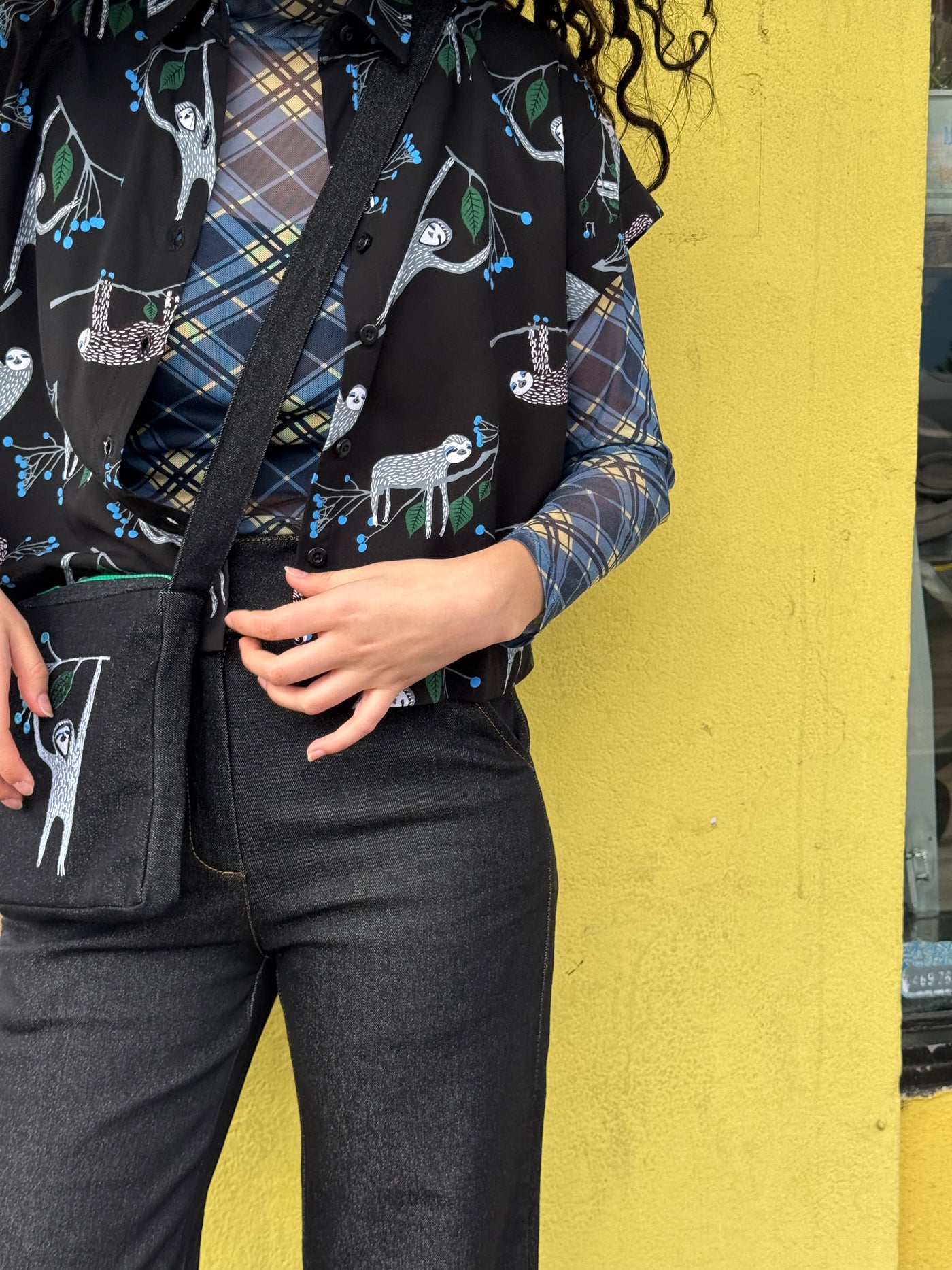 Person wearing a plaid shirt with sloth print shirt, matching bag and black pants against a yellow wall.