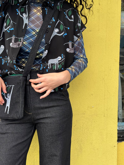 Person wearing a plaid shirt with sloth print shirt, matching bag and black pants against a yellow wall.