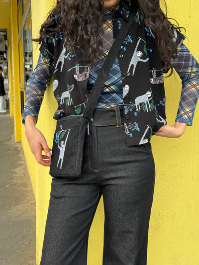 Person wearing a plaid shirt with sloth print shirt, matching bag and black pants against a yellow wall.