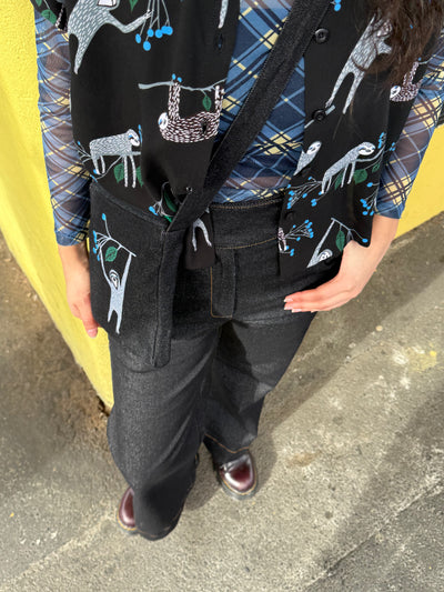 Person wearing a plaid shirt with sloth print shirt, matching bag and black pants against a yellow wall.