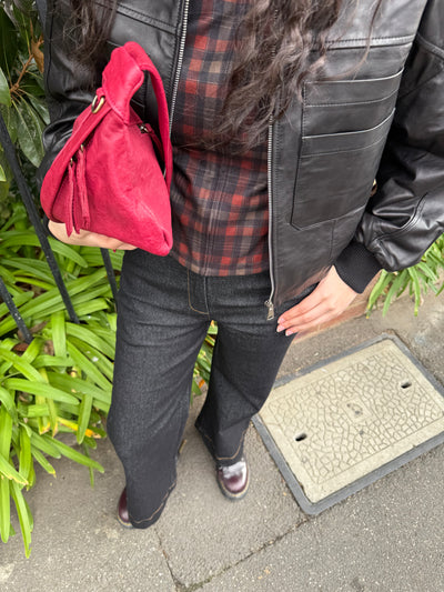 Person holding a red leather bag and wearing a black leather jacket over a plaid shirt with black jeans standing on a street with greenery