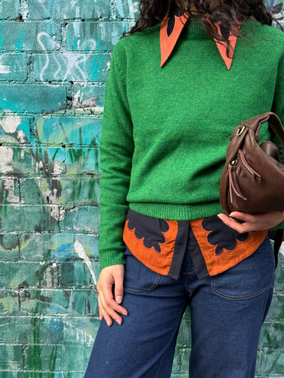 Person wearing a green sweater with a collared shirt under with orange and black patterns against a graffiti-covered wall.