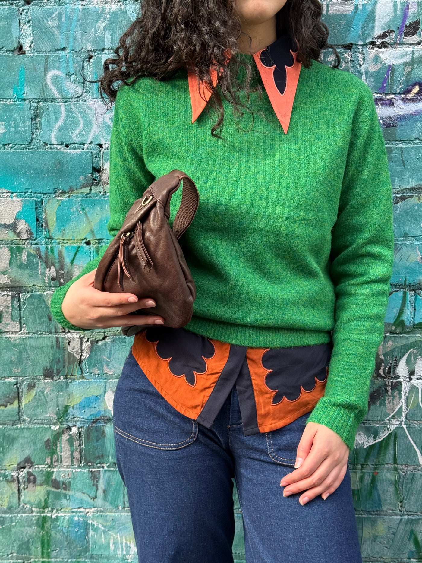 Person wearing a green sweater with a collared shirt under with orange and black patterns against a graffiti-covered wall.
