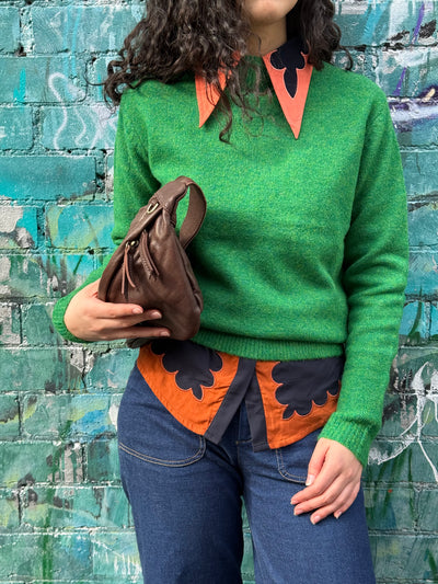 Person wearing a green sweater with a collared shirt under with orange and black patterns against a graffiti-covered wall.