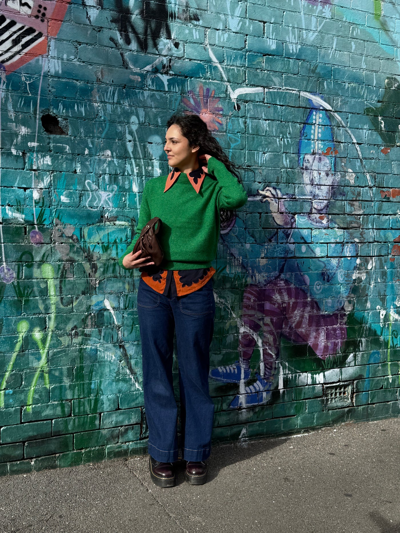 Woman in a green sweater and blue jeans standing against a graffiti-covered wall.