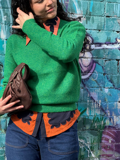 Person wearing a green sweater over an orange and black patterned shirt against a graffiti-covered wall.