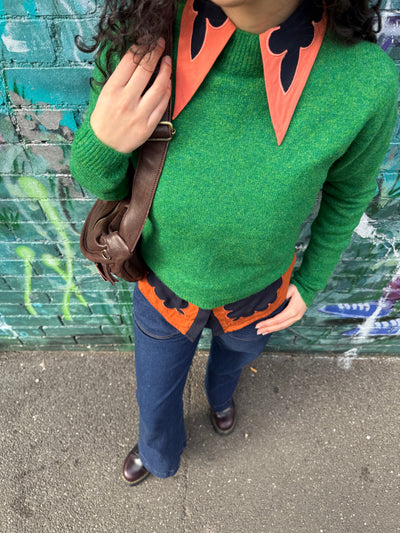 Person wearing a green sweater with orange and black accents, standing against a graffiti-covered wall.