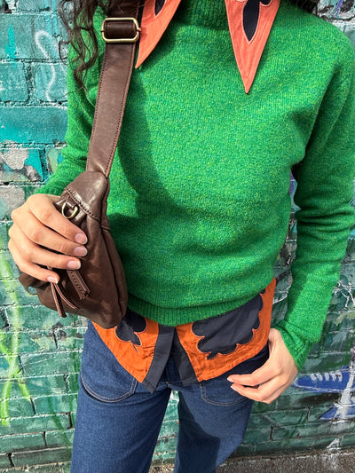 Person wearing a green sweater with orange accents and a brown bag against a graffiti-covered wall.