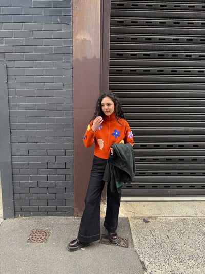 Woman wearing an orange sweater with colorful patterns and black flare jeans, standing against a gray wall.