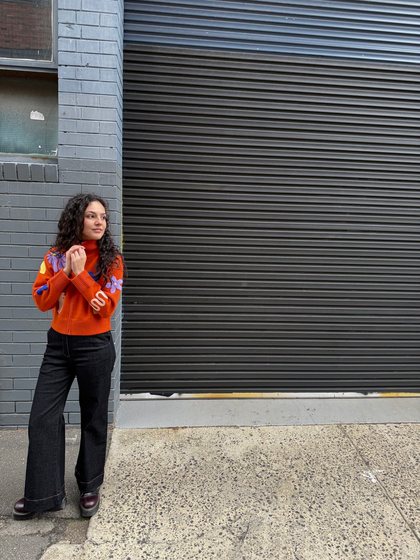 Person wearing an orange sweater and black jeans standing against a gray wall.