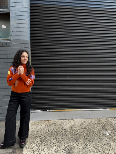 Woman standing against a closed metal shutter on a street wearing an orange cardigan and black flare jeans