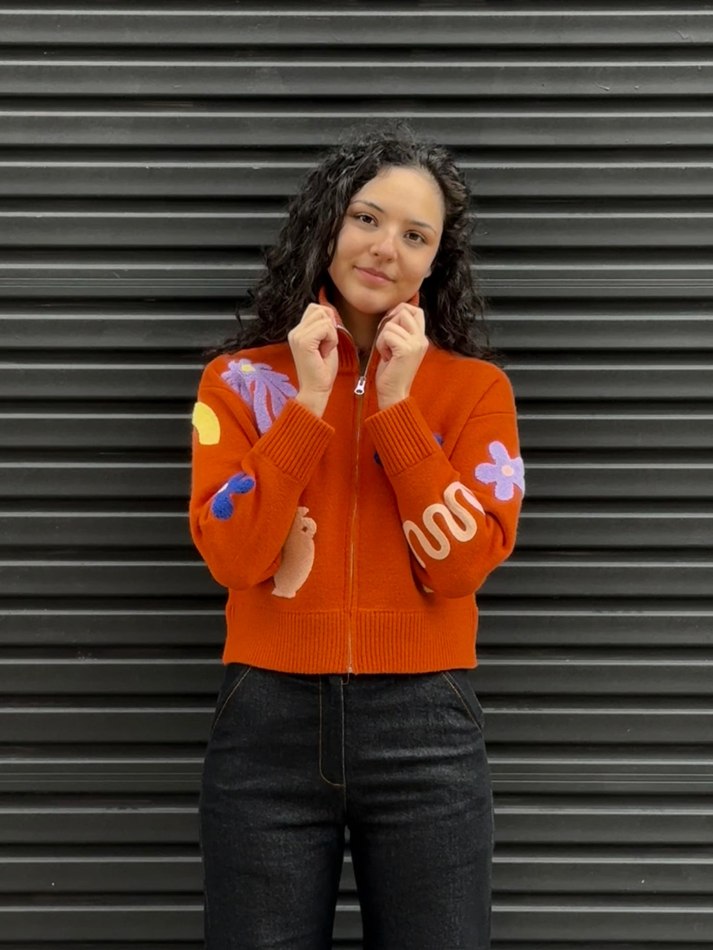 Person wearing an orange sweater with colorful designs and black jeans against a gray background