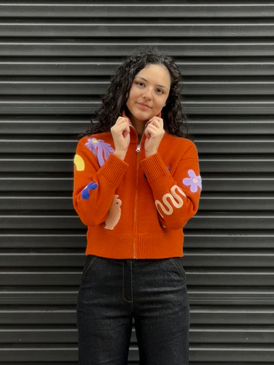 Person wearing an orange sweater with colorful designs and black jeans against a gray background